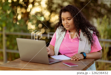 A young woman with curly hair engages with her laptop at a wooden table. She enjoys a productive moment outdoors, immersed in her work while surrounded by nature. A young woman with curly hair engages with her laptop at a wooden table. She enjoys a productive moment outdoors, immersed in her work while surrounded by nature. 126236432