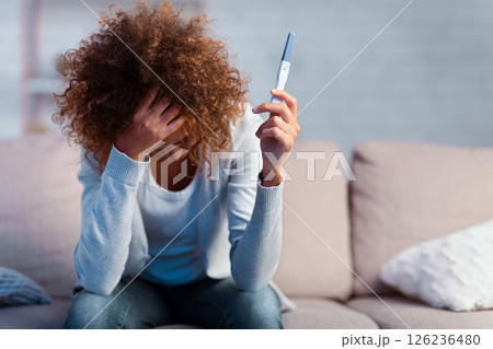 Depressed girl holding pregnancy test covering face with hand, sitting on couch, free space 126236480