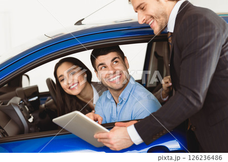 Check Out The Characteristics. Happy couple sitting in salon of new car, vendor showing tablet to them Check Out The Characteristics. Happy couple sitting in salon of new car, vendor showing tablet to them 126236486