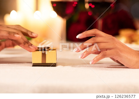 Romantic Proposal Concept. Closeup Of Male Hands Pushing Wrapped Gift Ring Box To Girlfriend's Hand On Served Restaurant Table. Cropped Romantic Proposal Concept. Closeup Of Male Hands Pushing Wrapped Gift Ring Box To Girlfriend's Hand On Served Restaurant Table. Cropped 126236577