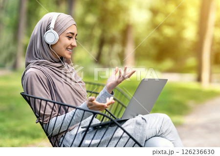 Pretty muslim girl having video conference while resting at park, using laptop and headset, empty space Pretty muslim girl having video conference while resting at park, using laptop and headset, empty space 126236630