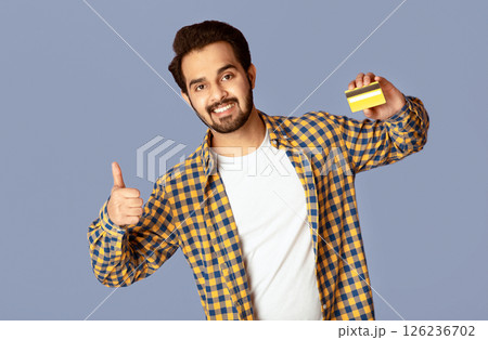Happy Indian guy holding credit card and showing thumb up gesture on violet background 126236702