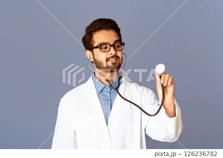 Young Indian doctor in lab coat using stethoscope on lilac background, copy space Young Indian doctor in lab coat using stethoscope on lilac background, copy space 126236782