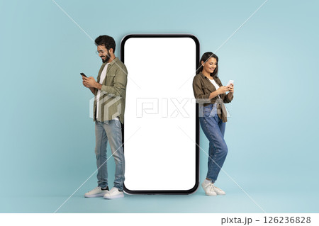 Stylish beautiful young eastern couple posing by huge phone with white blank screen, cheerful handsome indian man and pretty long-haired woman using smartphones, colorful background, mockup 126236828