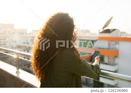 back view, curly girl stands in apartment on balloon with city view, holds phone in hands at sunset, communication, correspondence, online shopping, remote work, freelancer back view, curly girl stands in apartment on balloon with city view, holds phone in hands at sunset, communication, correspondence, online shopping, remote work, freelancer 126236960