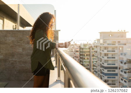 Young curly woman standing on balcony overlooking city at sunset with phone in hands, online shopping, social networking, remote work, side view Young curly woman standing on balcony overlooking city at sunset with phone in hands, online shopping, social networking, remote work, side view 126237082