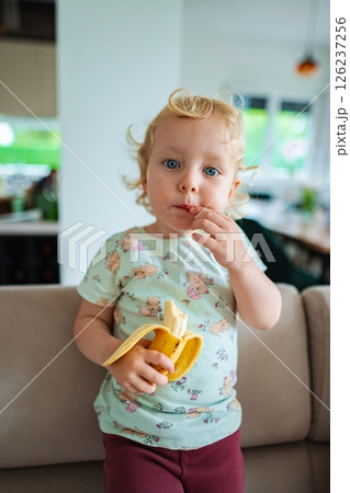 Curious toddler enjoys banana snack in cozy home setting Curious toddler enjoys banana snack in cozy home setting 126237256