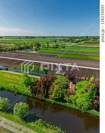 Aerial view of a lush countryside garden surrounded by canals and farmlands, with a cozy house and colorful trees, set in a peaceful rural landscape under a clear blue sky. 126238895