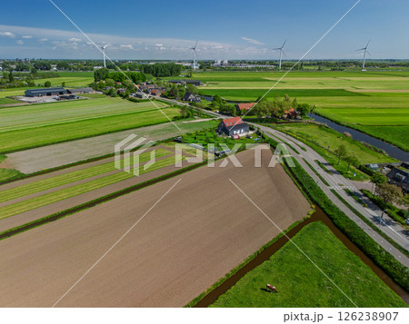 Aerial view of green farmlands with grazing cows, freshly plowed fields, and distant wind turbines under a clear blue sky in the Dutch countryside. 126238907