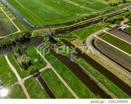 Aerial view of a Dutch countryside with a traditional windmill by a canal, green fields, and a nearby road with moving cars under a clear blue sky. 126238908