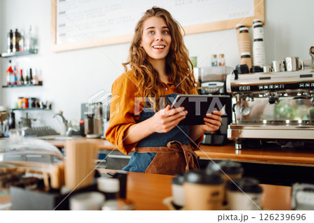Smiling female barista takes an order from a tablet while standing at bar counter in a coffee shop. Smiling female barista takes an order from a tablet while standing at bar counter in a coffee shop. 126239696