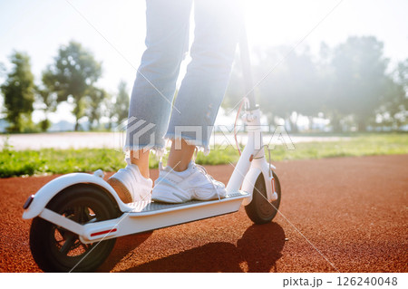Woman riding electric kick scooter outdoors at sunset, closeup. Ecological transportation concept. Woman riding electric kick scooter outdoors at sunset, closeup. Ecological transportation concept. 126240048