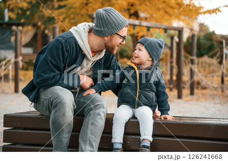 Sitting on the bench and having fun. Father and young son is together outdoors at daytime Sitting on the bench and having fun. Father and young son is together outdoors at daytime 126241668