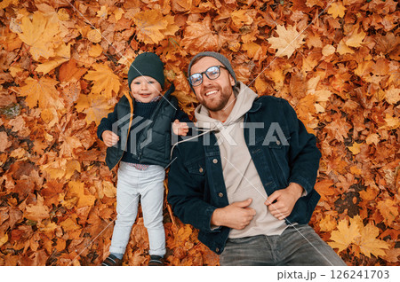 Laying down on the ground that covered with fallen leaves. Father and young son is together outdoors at daytime Laying down on the ground that covered with fallen leaves. Father and young son is together outdoors at daytime 126241703