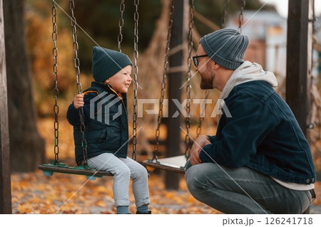 Boy sits on a swing. Father and young son is together outdoors at daytime Boy sits on a swing. Father and young son is together outdoors at daytime 126241718