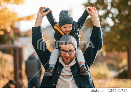 Front view. Son is on the dad's shoulders. Father and young son is together outdoors at daytime Front view. Son is on the dad's shoulders. Father and young son is together outdoors at daytime 126241722