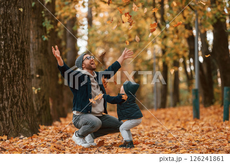 Playing with fallen leaves. Father and young son is together outdoors at daytime Playing with fallen leaves. Father and young son is together outdoors at daytime 126241861