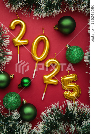 Merry Christmas and Happy New Year 2025. Golden candles numbers 2025 with green balls on red background. Top view 126242596