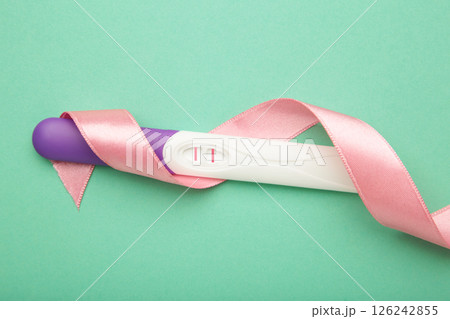 The pregnancy test with two stripes with pink ribbon on mint background. Health care concept. Top view The pregnancy test with two stripes with pink ribbon on mint background. Health care concept. Top view 126242855