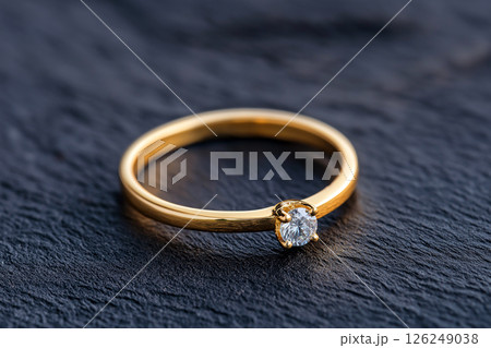 Elegant golden ring featuring a single brilliant cut diamond, placed on a dark textured surface. Symbolizing love, commitment, and luxury, this piece highlights the sophistication of fine jewelry. 126249038
