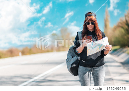 Caucasian woman in cap and sunglasses stand along the road with a paper map and using a smartphone. Copy space. Concept of online guide and hitchhiking 126249986