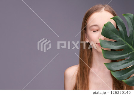 Young caucasian female with long hair and monstera leaf on gray background Young caucasian female with long hair and monstera leaf on gray background 126250140