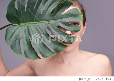 Young caucasian female partially concealed by monstera leaf against neutral 126250193