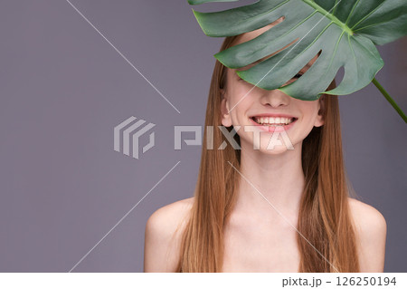 Smiling young caucasian female partially hidden by monstera leaf against purple 126250194