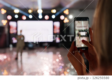 defocus. A woman is filming a man's performance on stage on her smartphone. defocus. A woman is filming a man's performance on stage on her smartphone. 126250380