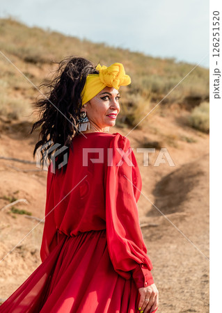 A woman in a red dress and yellow headband is walking on a dirt path. She is smiling and she is happy. 126251520