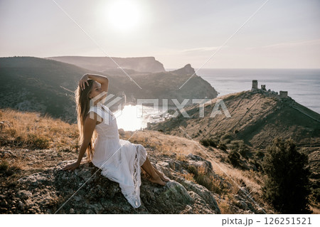 A woman in a white dress is sitting on a rock overlooking a body of water. She is enjoying the view and taking in the scenery. 126251521