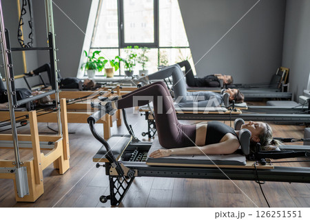 Group of caucasian women exercising with trainer on pilates reformers in a bright studio. Their workout focuses on core strength and flexibility in a modern fitness environment Group of caucasian women exercising with trainer on pilates reformers in a bright studio. Their workout focuses on core strength and flexibility in a modern fitness environment 126251551