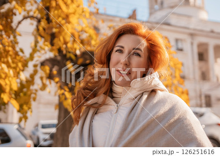Woman Autumn Portrait Outdoors: Redhead posing happily near yellow leaves, city background, showcasing fall fashion. 126251616