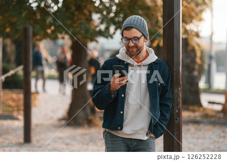 Standing with smartphone. Beautiful man in warm clothes is outdoors in the park Standing with smartphone. Beautiful man in warm clothes is outdoors in the park 126252228