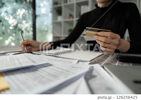 Woman holding a credit card while reviewing financial papers and taking notes at home. Woman holding a credit card while reviewing financial papers and taking notes at home. 126256425
