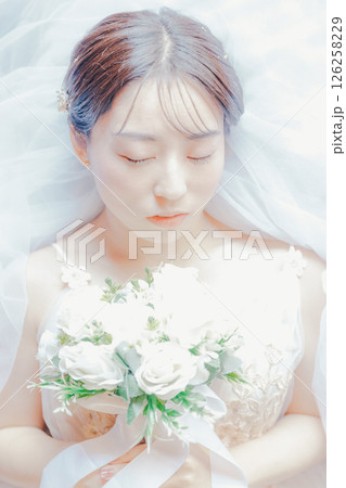 純白のドレスをまとい、ブーケを胸元に持って横たわる女性のイメージ写真 126258229