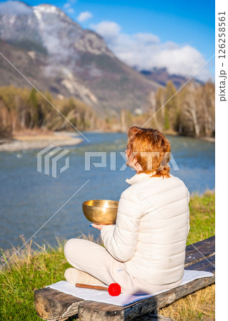 Woman playing a Tibetan singing bowl by a mountain river in Altai. Concept of sound healing, spiritual alignment, and deep connection with natural elements 126258561