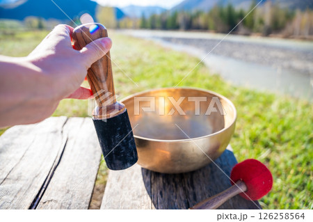 Point of view of hand playing a Tibetan singing bowl near mountain river in Altai. Concept of personal ritual, sound healing, and spiritual harmony with wild nature. 126258564