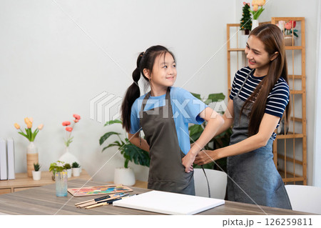 Creative bonding through art: Mother helping daughter wear an apron Creative bonding through art: Mother helping daughter wear an apron 126259811
