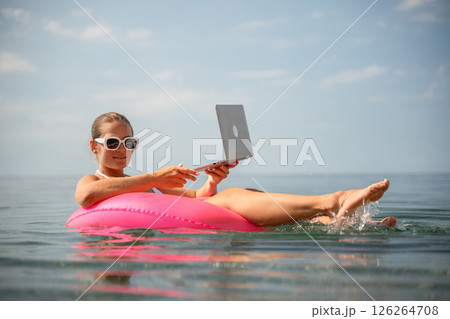 A woman is floating on a pink inflatable raft in the ocean while holding a laptop. Concept of relaxation and leisure, as the woman is enjoying her time in the water while working on her laptop. A woman is floating on a pink inflatable raft in the ocean while holding a laptop. Concept of relaxation and leisure, as the woman is enjoying her time in the water while working on her laptop. 126264708