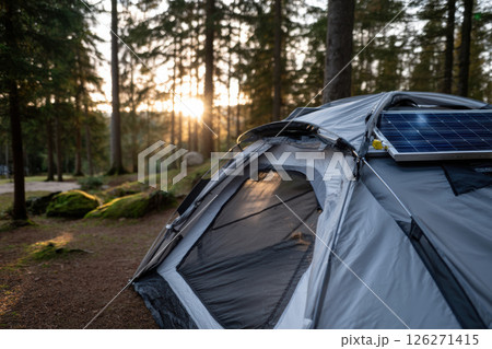 Tent equipped with solar panel is set up in serene forest, capturing warm glow of sunset filtering through trees. peaceful setting highlights sustainable camping practices Tent equipped with solar panel is set up in serene forest, capturing warm glow of sunset filtering through trees. peaceful setting highlights sustainable camping practices 126271415