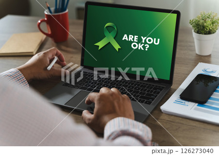 Mental Health Awareness person holding pen front of laptop with green awareness ribbon and text ARE YOU OK screen, surrounded by office supplies 126273040