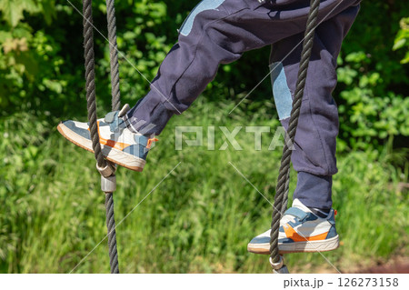 Child in sports shoes climbing a rope ladder at an outdoor playground. The image shows focus on the child's legs and shoes, highlighting activity and movement. 126273158