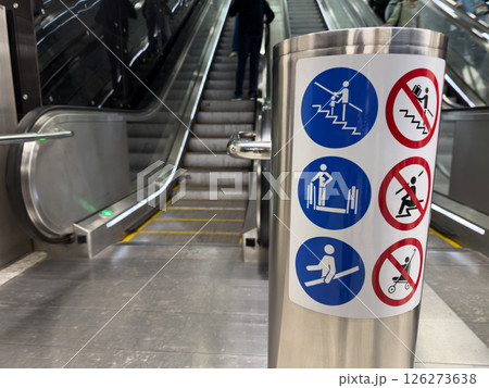 Closeup of escalator safety instruction sign in metro station with visible stairs and people in background, illustrating public space design and visual safety communication Closeup of escalator safety instruction sign in metro station with visible stairs and people in background, illustrating public space design and visual safety communication 126273638