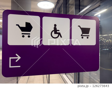 Purple directional sign with black icons indicating areas for baby strollers, wheelchair users and shopping carts, placed in modern parking facility with glass walls and lighting reflections Purple directional sign with black icons indicating areas for baby strollers, wheelchair users and shopping carts, placed in modern parking facility with glass walls and lighting reflections 126273643
