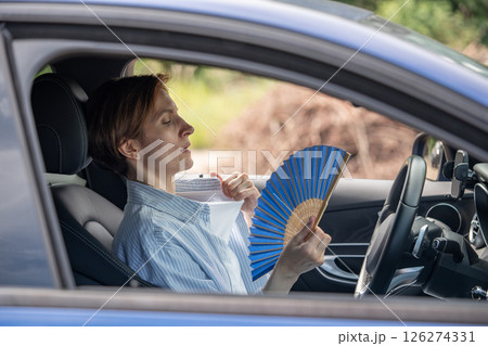Tired exhausted middle aged woman waving blue fan suffers from stuffiness driving car.  126274331