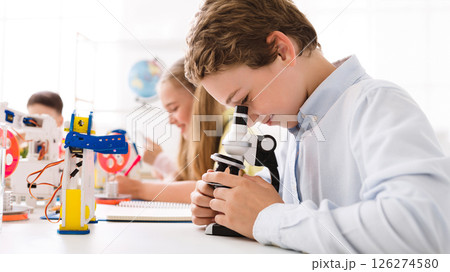 A young boy, wearing a white shirt, looks intently through a microscope, focusing on a slide. He is sitting at a desk in a classroom setting, surrounded by other students and educational materials A young boy, wearing a white shirt, looks intently through a microscope, focusing on a slide. He is sitting at a desk in a classroom setting, surrounded by other students and educational materials 126274580