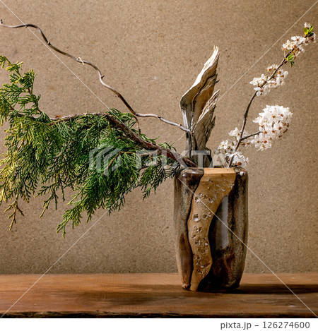 Beautiful spring ikebana. Floral arrangement with spring blooming white flowers, thuja branch and piece of wood in brown ceramic vase, standing on wooden table. Japanese style home decor Beautiful spring ikebana. Floral arrangement with spring blooming white flowers, thuja branch and piece of wood in brown ceramic vase, standing on wooden table. Japanese style home decor 126274600