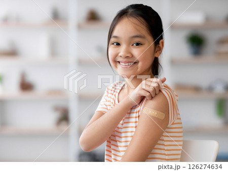 Healthy asian kid girl got vaccinated, sitting on couch at home and showing adhesive band on her shoulder, smiling at camera, panorama with copy space. Kids vaccination, immunization concept 126274634