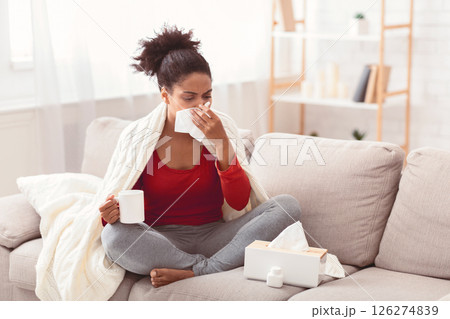 Seasonal Flu. Sick Afro Woman Blowing Nose In Paper Tissue Feeling Bad Sitting On Sofa At Home. Copy Space 126274839